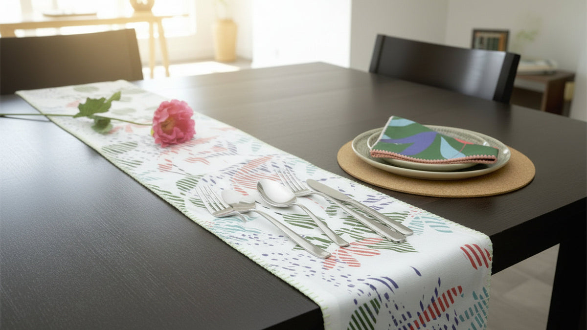 A white table runner with a colorful botanical and abstract pattern is styled on a dark wooden dining table. The runner is decorated with silver flatware, a pink rose stem, and a matching patterned napkin on a round cork placemat from Hem And Tassel 