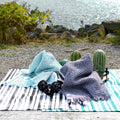 Two textured waffle kitchen towels, one blue and one purple, on a striped kitchen towel with cacti in a natural setting from Hem And Tassel