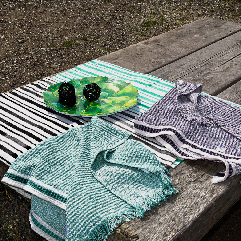 Textiles including a green towel, striped fabric, and a small dish on a wooden surface from Hem And Tassel 