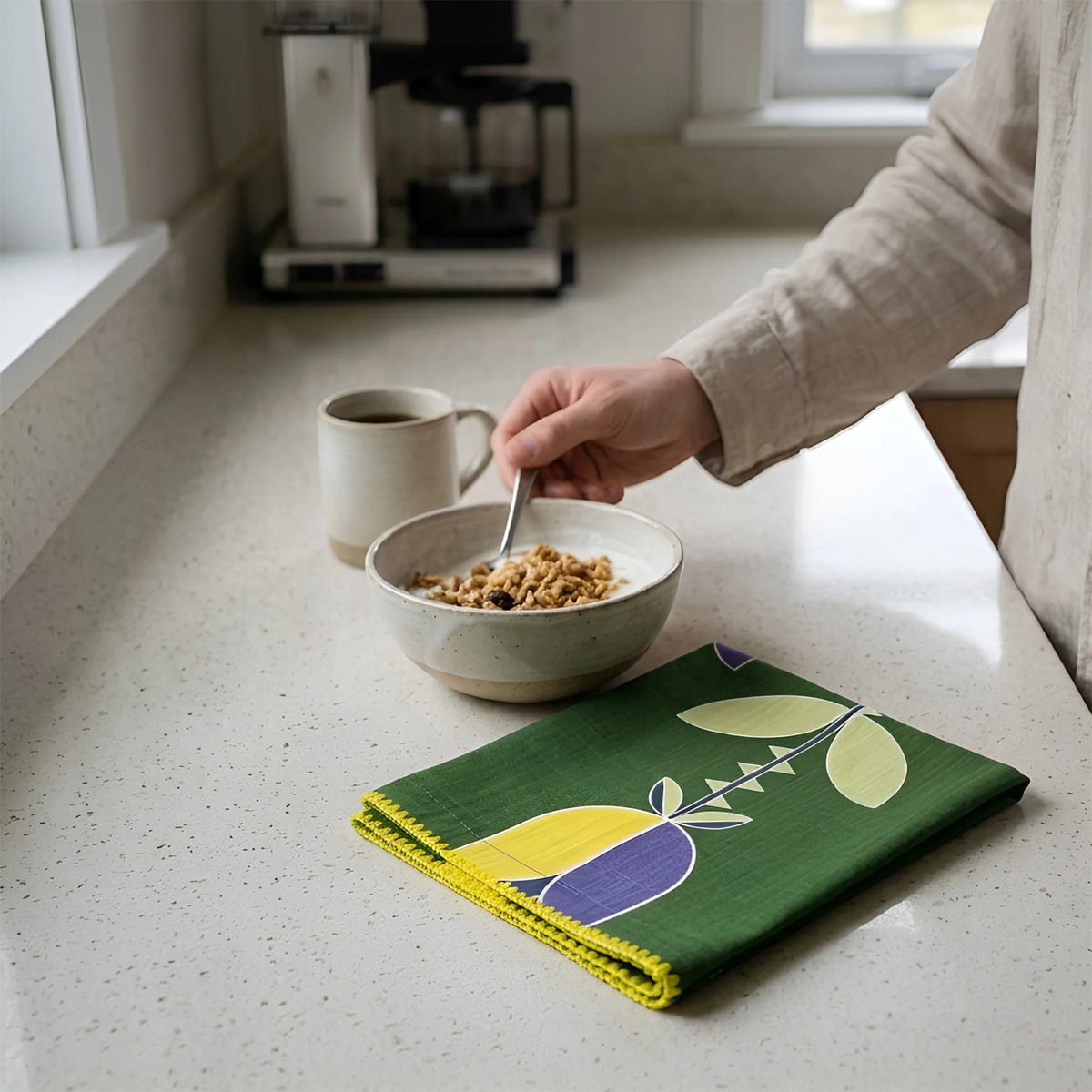 A green cotton textured napkin with a yellow and purple tulip pattern from Hem And Tassel is folded on a speckled white countertop next to a bowl of cereal and a mug, with a person's hand reaching for the cereal.