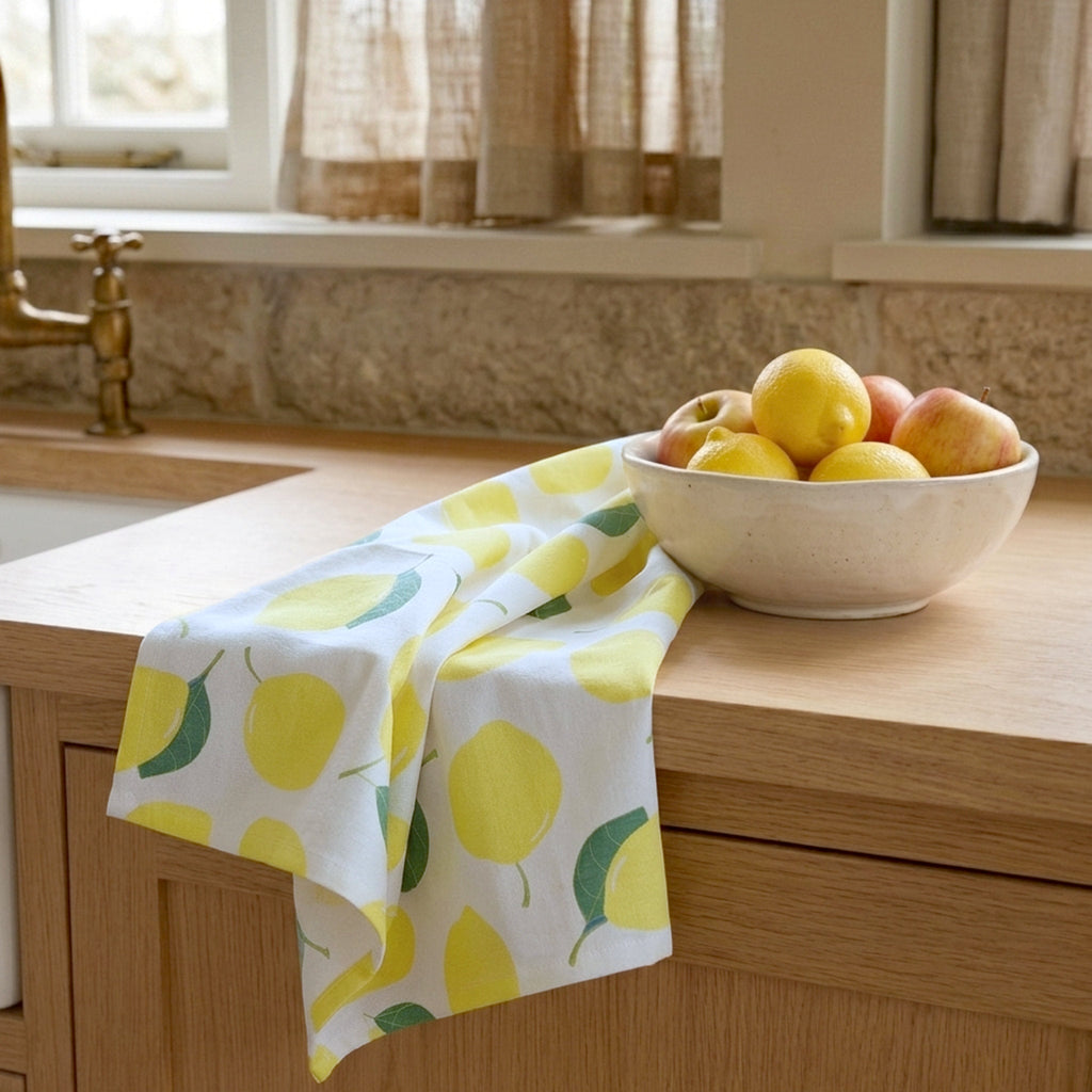 A white kitchen towel with a yellow lemon and green leaf pattern draped over a wooden kitchen counter next to a bowl of fruit from Hem And Tassel