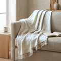 A cream-colored throw blanket with a beige floral damask pattern and fringed edges, draped over a beige fabric sofa in a cozy, sunlit living room setting from Hem And Tassel 