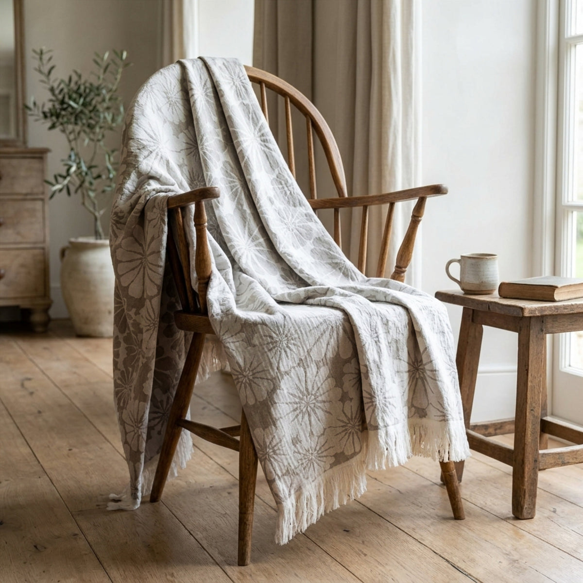 A light slate and white patterned throw blanket with fringe is draped over a wooden spindle-back chair in a bright, rustic room with a side table and window from Hem And Tassel 