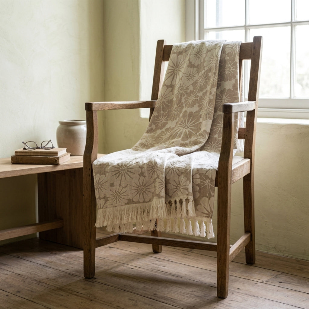 A slate and beige patterned fringed throw blanket draped over a wooden armchair in a softly lit room with a window, side table, and ceramic vase.