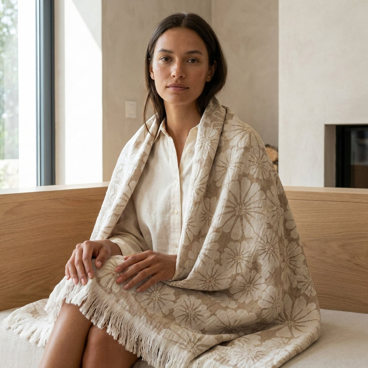 A woman sits on a wooden bench wrapped in a large slate and beige patterned throw blanket with fringe, in a softly lit indoor setting from Hem And Tassel 