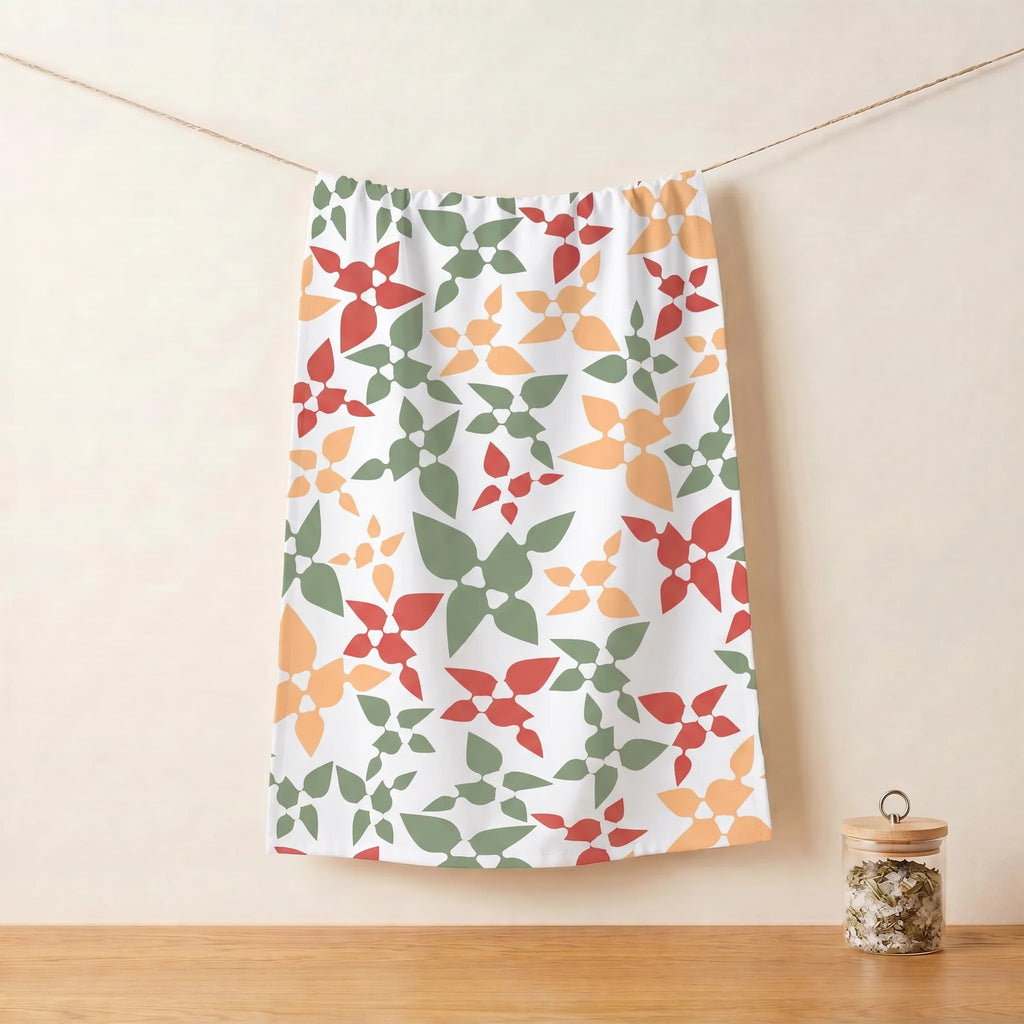 A white cotton kitchen towel with a repeating geometric floral pattern in green, orange, and red, hanging from a string against a beige wall with a wooden surface below from Hem And Tassel