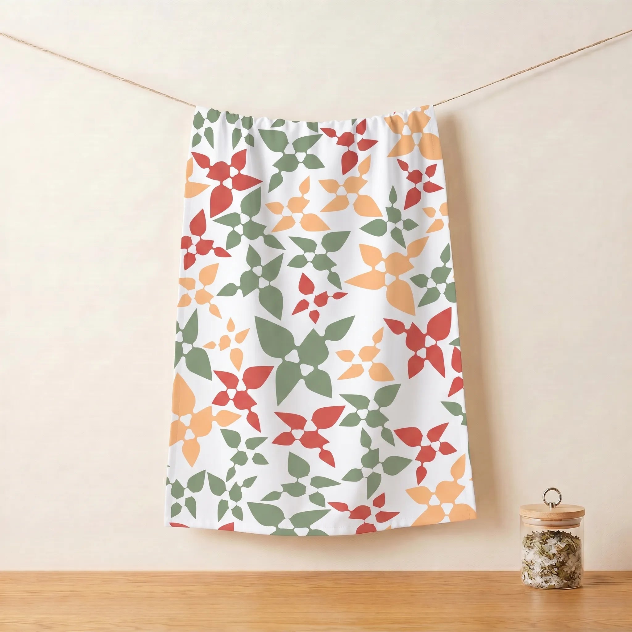A white cotton kitchen towel with a repeating geometric floral pattern in green, orange, and red, hanging from a string against a beige wall with a wooden surface below from Hem And Tassel