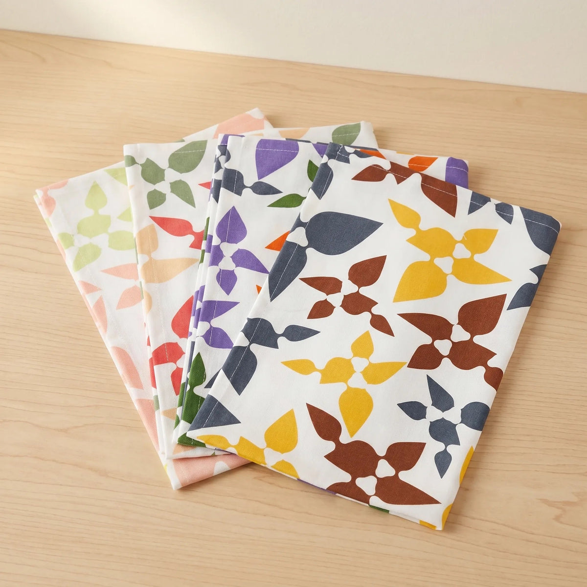 Four folded cotton kitchen towels with a retro geometric leaf pattern in various colors, arranged in a fan shape on a light wood tabletop from Hem And Tassel
