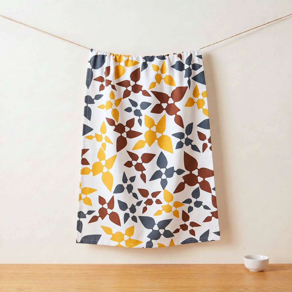 A white cotton kitchen towel with a retro geometric floral pattern in mustard yellow, terracotta brown, and slate blue, hanging from a thin cord against a warm off-white wall with a wooden tabletop below from Hem And Tassel