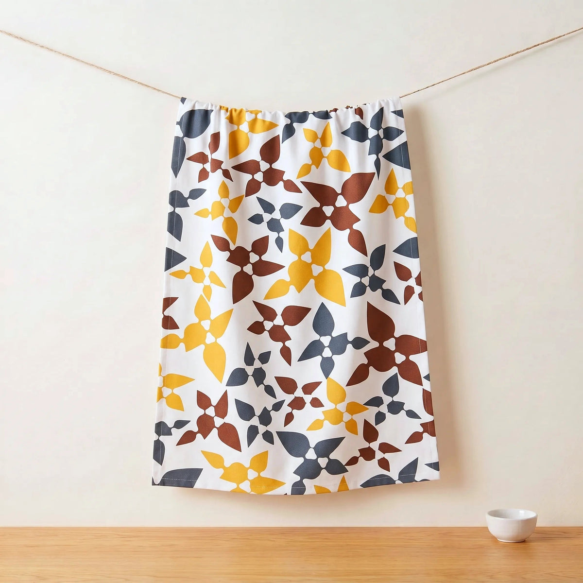 A white cotton kitchen towel with a retro geometric floral pattern in mustard yellow, terracotta brown, and slate blue, hanging from a thin cord against a warm off-white wall with a wooden tabletop below from Hem And Tassel