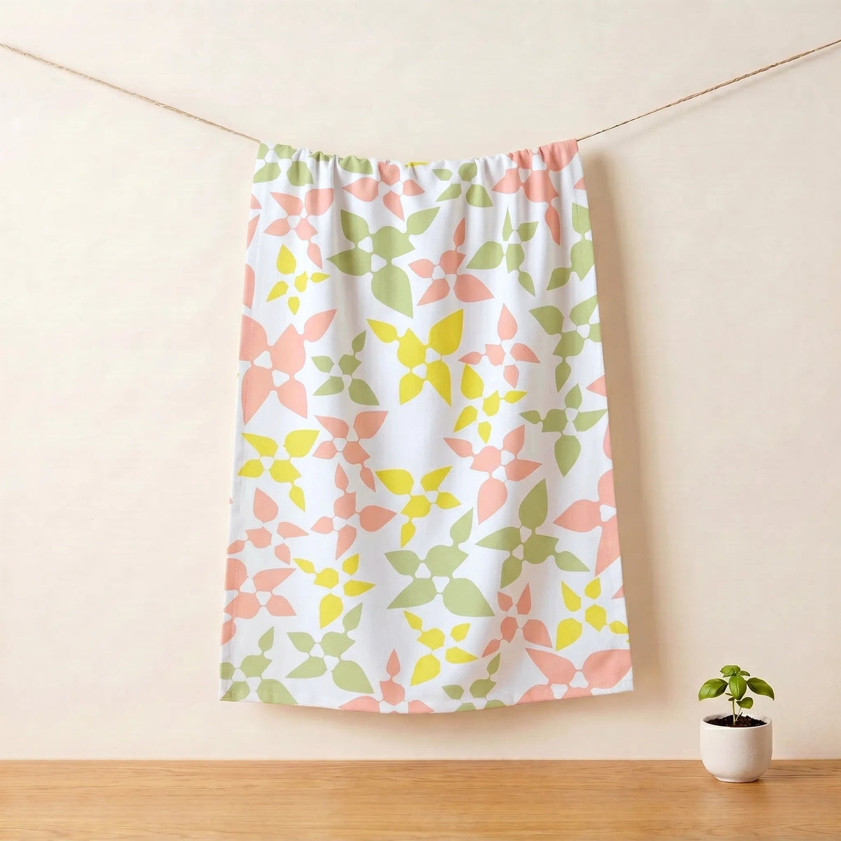 A cotton kitchen towel with a repeating pastel floral pattern in pink, yellow, and green, hanging from a string on a light beige wall above a wooden surface with a small potted plant from Hem And Tassel