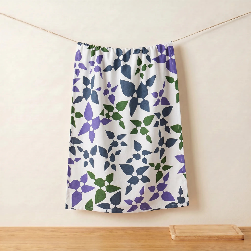 A white cotton kitchen towel  featuring a repeating geometric floral pattern in purple, navy blue, and green, hanging from a string against a beige wall from Hem And Tassel