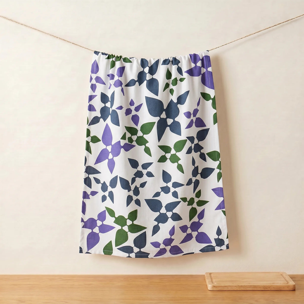 A white cotton kitchen towel  featuring a repeating geometric floral pattern in purple, navy blue, and green, hanging from a string against a beige wall from Hem And Tassel