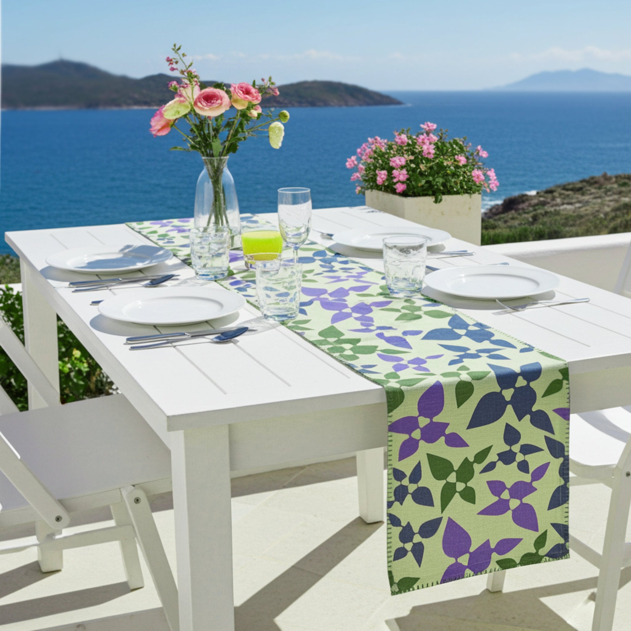 Outdoor table setting with a Celebration table runner, plates, glasses, and flowers against a scenic ocean backdrop from Hem And Tassel 