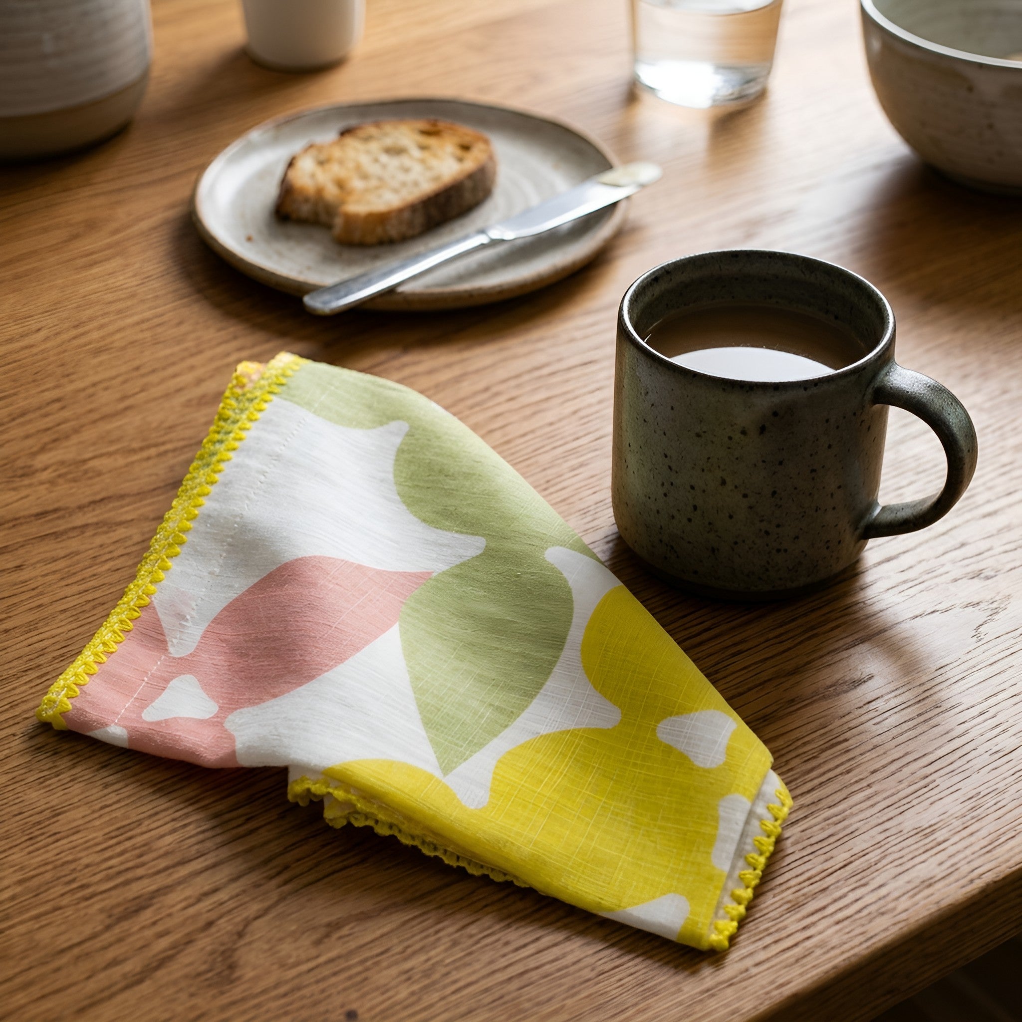 A folded patterned cloth napkin with yellow, pink, and green abstract shapes and yellow scalloped edges, placed on a wooden table beside a speckled gray ceramic mug filled with coffee. A plate with a slice of toast and a knife are blurred in the background from Hem And Tassel 
