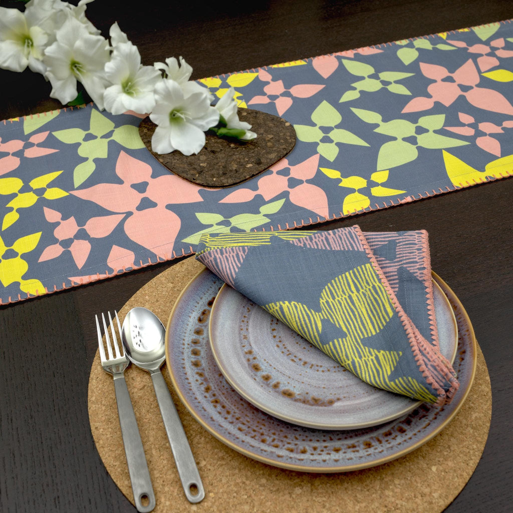 Table setting with floral-patterned Celebration dinner napkins and table runner, and cutlery on a dark surface from hem and tassel