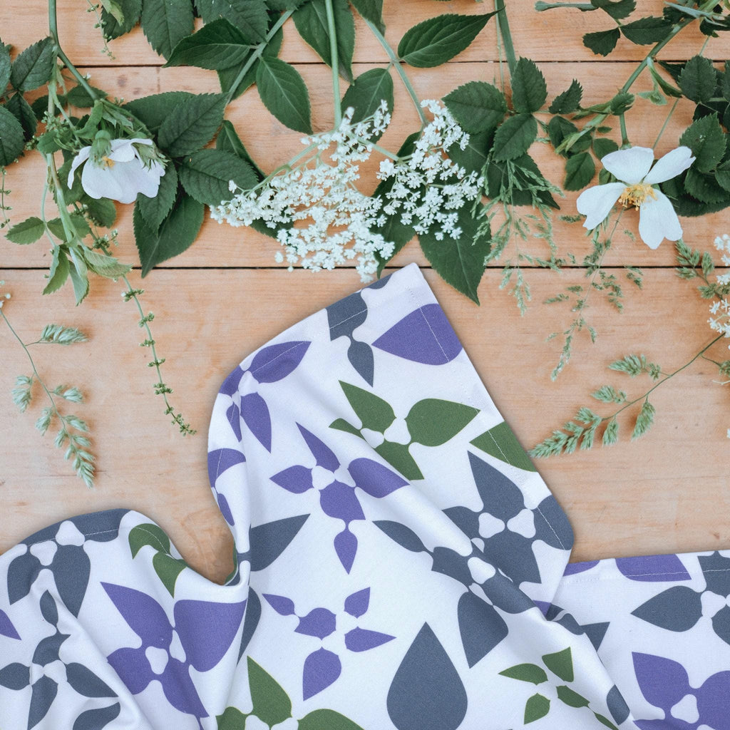 Celebration tea towel, with purple and green leaf pattern on a wooden surface with flowers and leaves from Hem And Tassel 