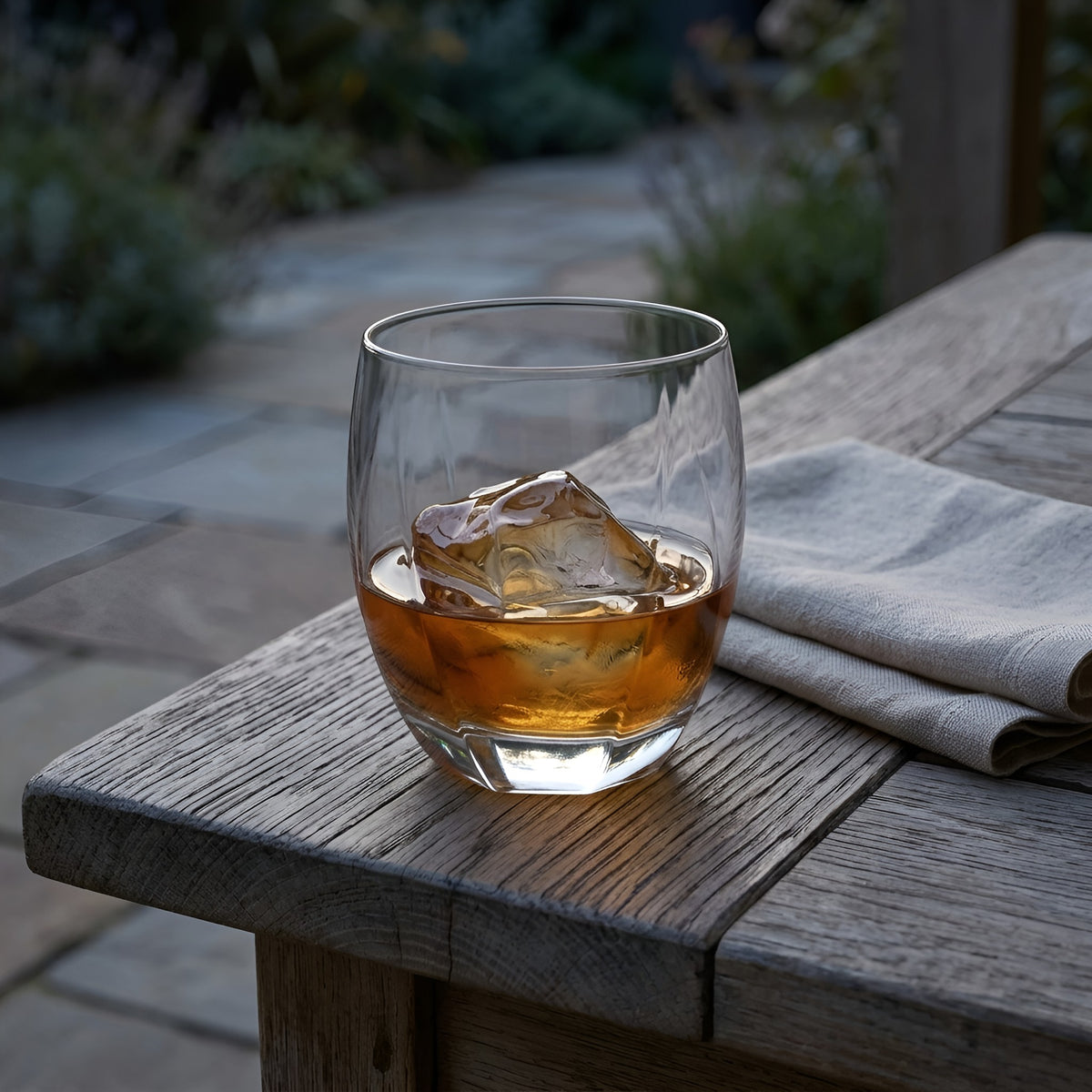 A clear lowball-style ridged drinking glass with a thick base, filled with amber liquid and a large ice cube, resting on a weathered wooden table outdoors with a folded napkin beside it from Hem And Tassel 