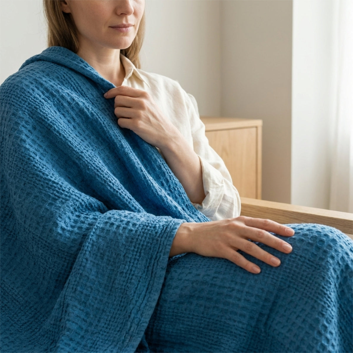 A woman wrapped in a complex waffle-knit blue throw blanket, wearing a cream shirt in a softly lit indoor setting from Hem And Tassel 