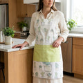 A woman wearing a white kitchen apron with a green and white cactus pattern and a solid light green front pocket, standing in a modern kitchen from Hem And Tassel 