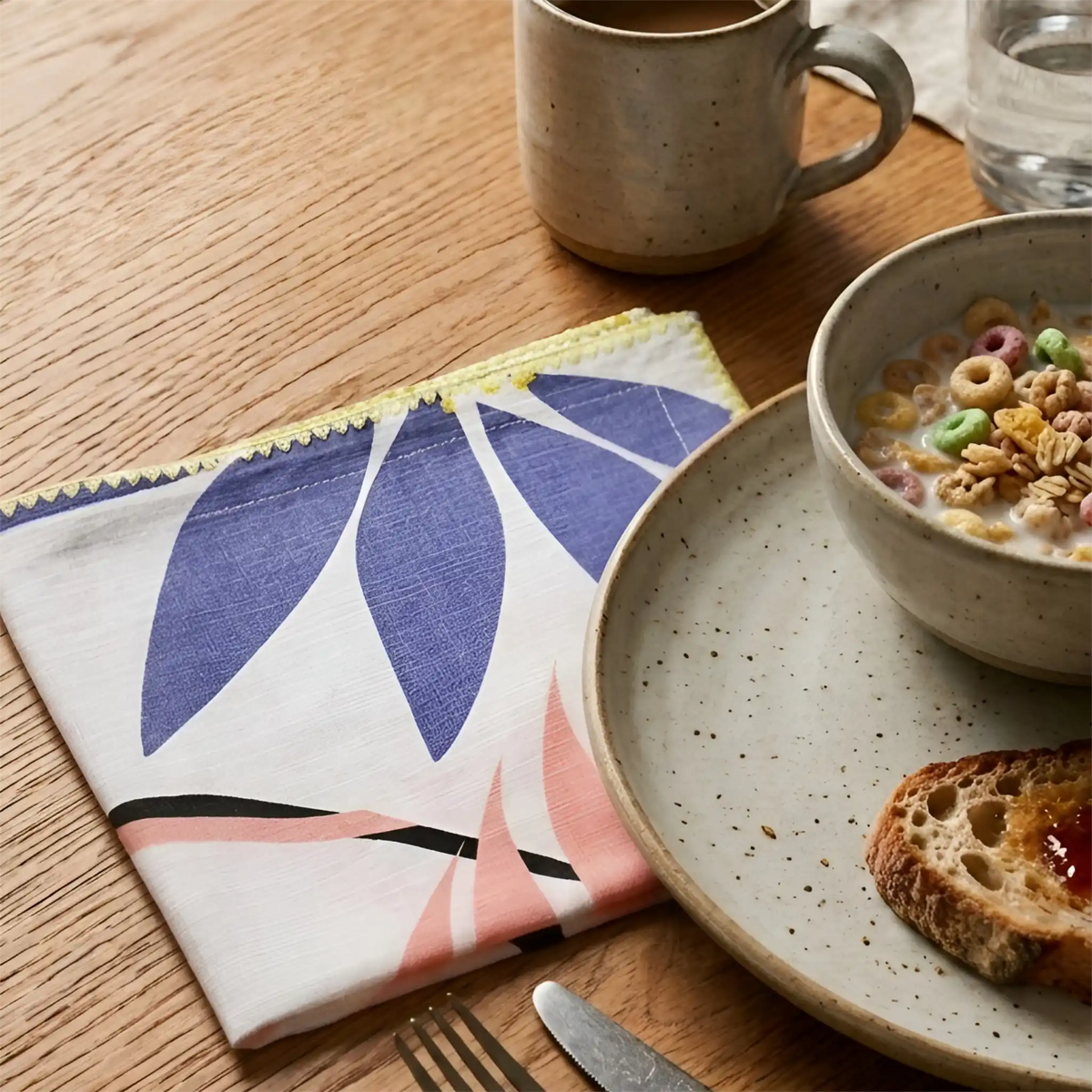 A folded patterned cotton textured napkin with blue, pink, yellow, and black leaf-like shapes on a white base, styled on a wooden table beside a ceramic mug, plate, bowl of cereal, and toast from Hem And Tassel 