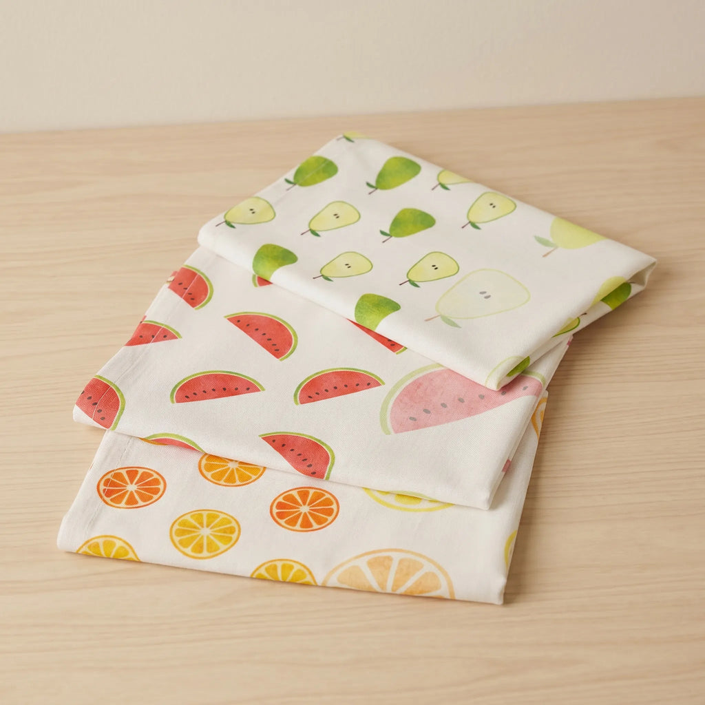 Three folded white cotton kitchen towels with fruit prints (pears, watermelons, oranges, and lemons) stacked on a light wood surface.