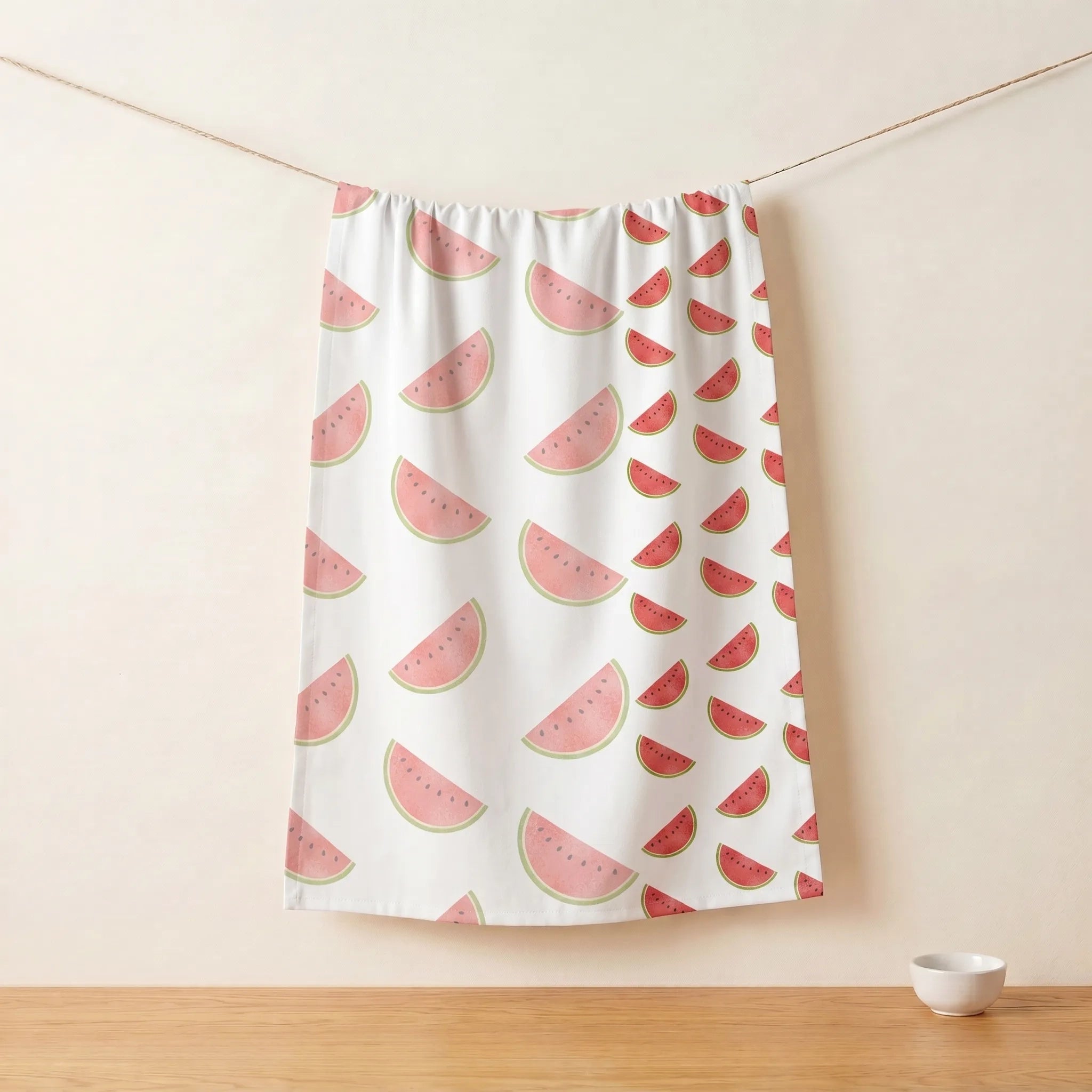A white pure cotton kitchen towel featuring a watermelon slice pattern, hanging from a string against a beige wall with a wooden tabletop and a small bowl below from Hem And Tassel