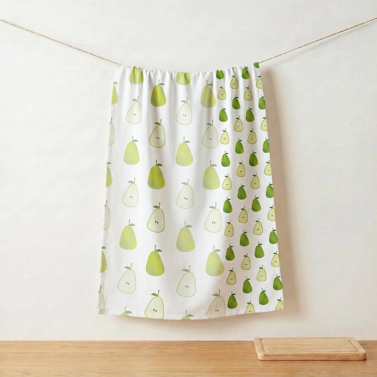 A white pure cotton kitchen towel featuring a pears' pattern, hanging from a string against a beige wall with a wooden tabletop and a small bowl below from Hem And Tassel