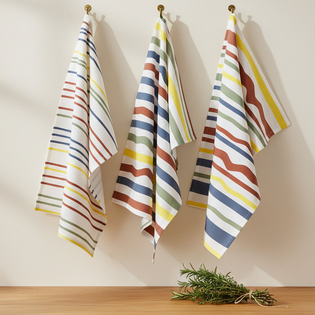 Fall  colors stripes in narrow, medium and broad widths on 3 cotton kitchen towels hanging on a beige wall with a sprig of rosemary  on a wood counter below from Hem And Tassel 