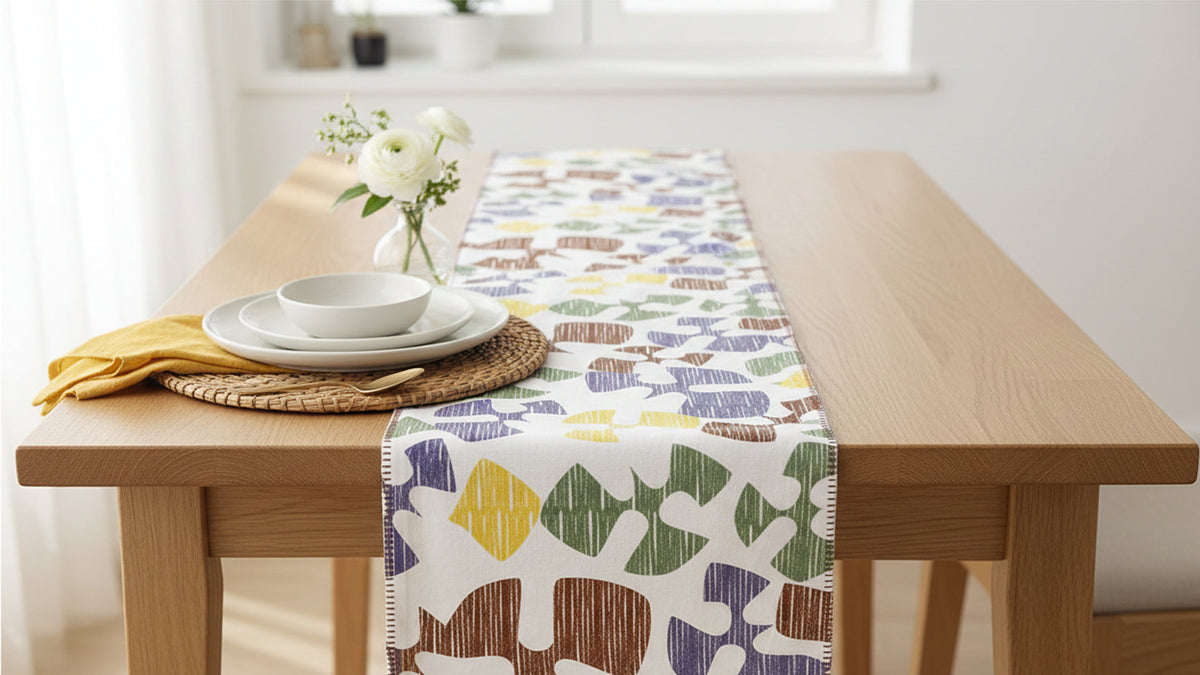 A patterned table runner with abstract shapes in purple, green, yellow, and brown on a white background, styled on a light wood dining table with a white bowl, plates, a yellow napkin, and a vase of white flowers from Hem And Tassel 