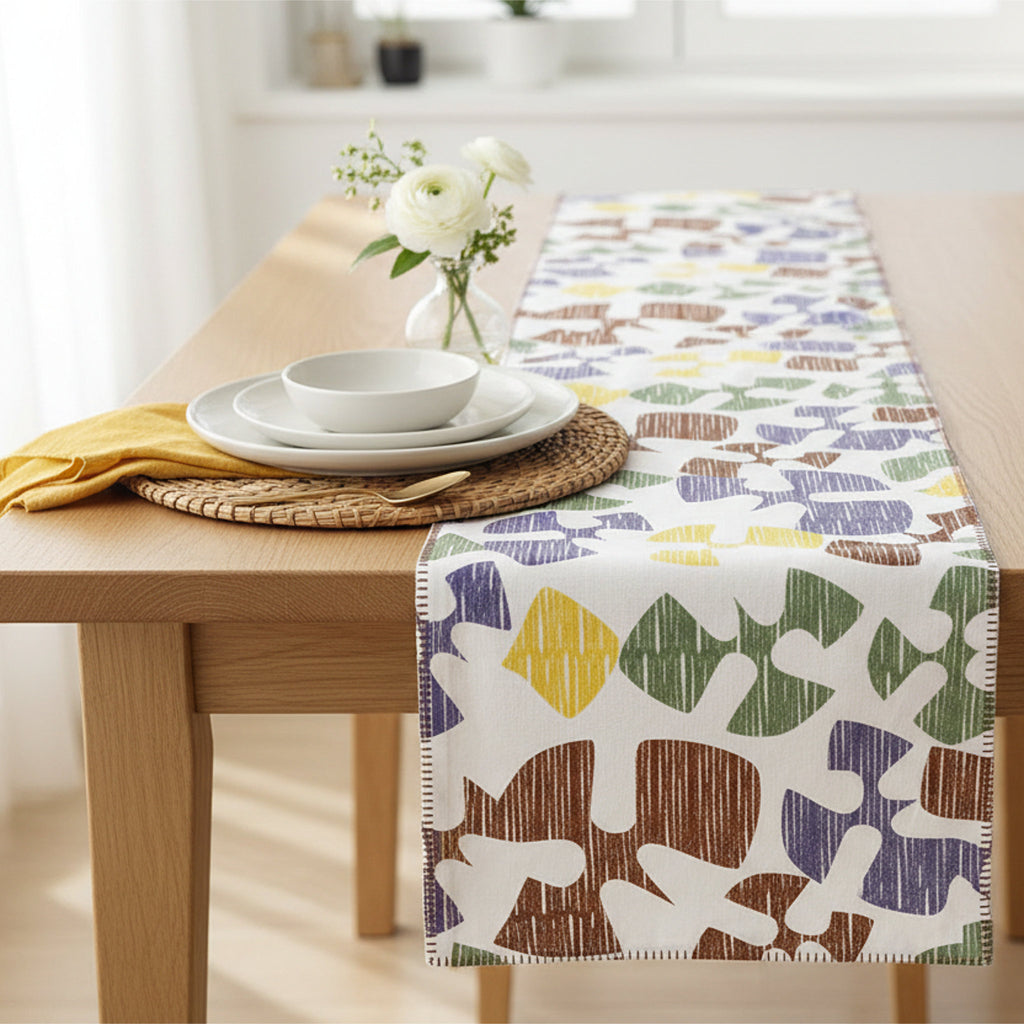 A patterned table runner draped over a light wood dining table, styled with white plates, a yellow napkin, and a small vase of white flowers in a bright room from Hem And Tassel 