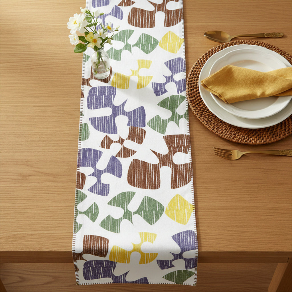 A table runner with a colorful abstract pattern of organic shapes in brown, green, yellow, and purple on a white background, styled on a light wood table with plates, a napkin, and a small vase of flowers from Hem And Tassel 
