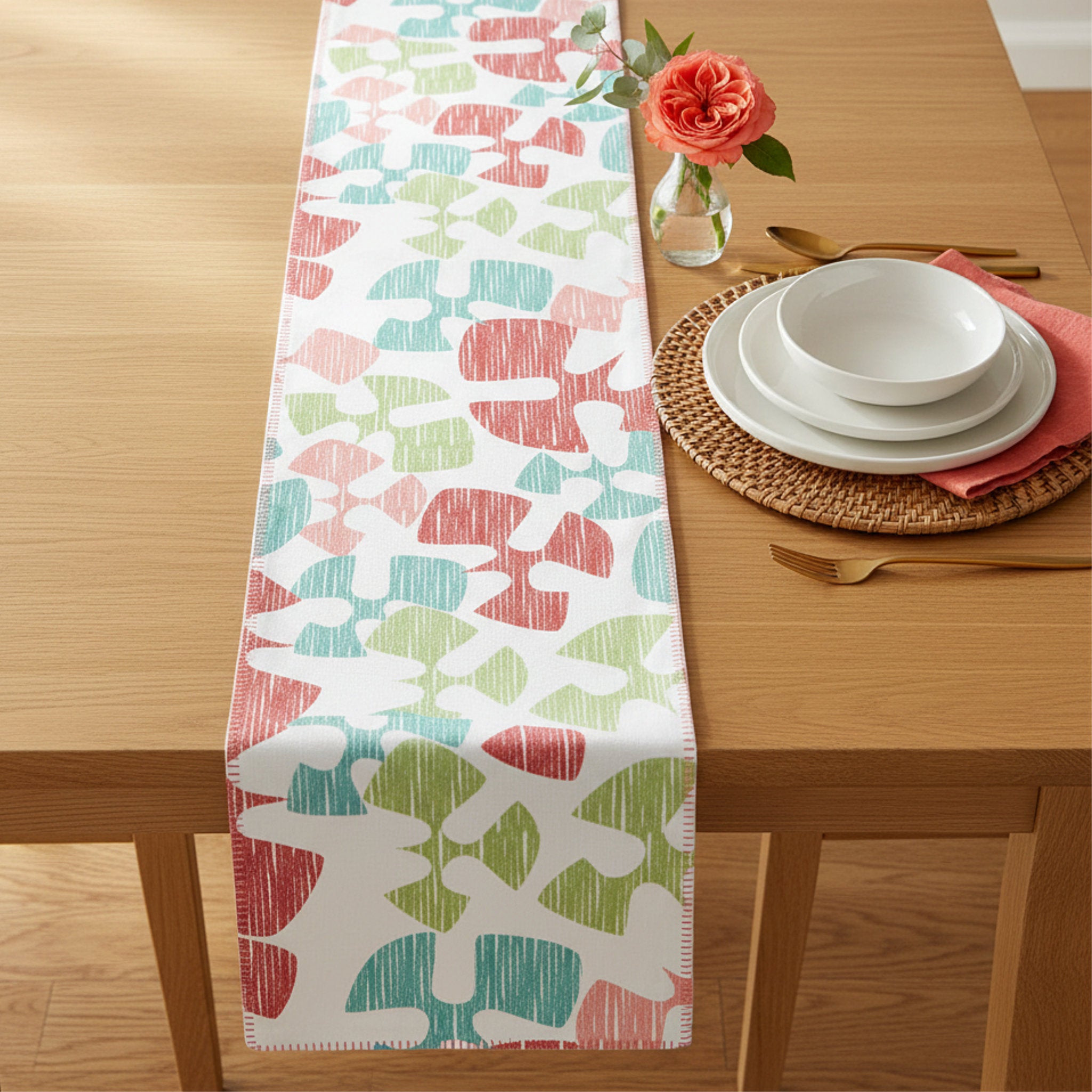 A rectangular table runner with a colorful abstract pattern in coral pink, teal, and light green on a white background, styled on a light wood dining table with a place setting and a vase of rose from Hem And Tassel 