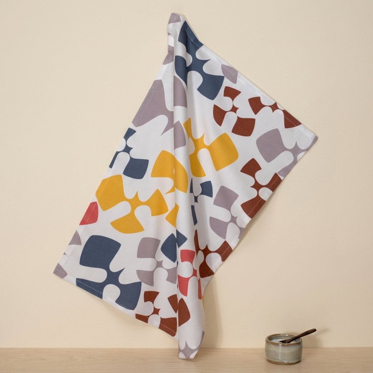A white cotton kitchen towel with a colorful pattern like abstract birds flying hangs from a hook on a beige wall above a wooden table with a small ceramic bowl and spoon from Hem And Tassel