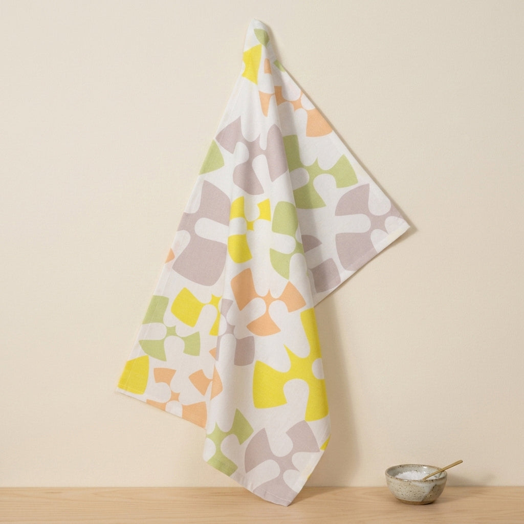 A white kitchen towel with a colorful abstract organic pattern of flying birds in yellow, orange, green, and gray, hanging on a wall hook next to a small ceramic bowl on a wooden surface from Hem And Tassel