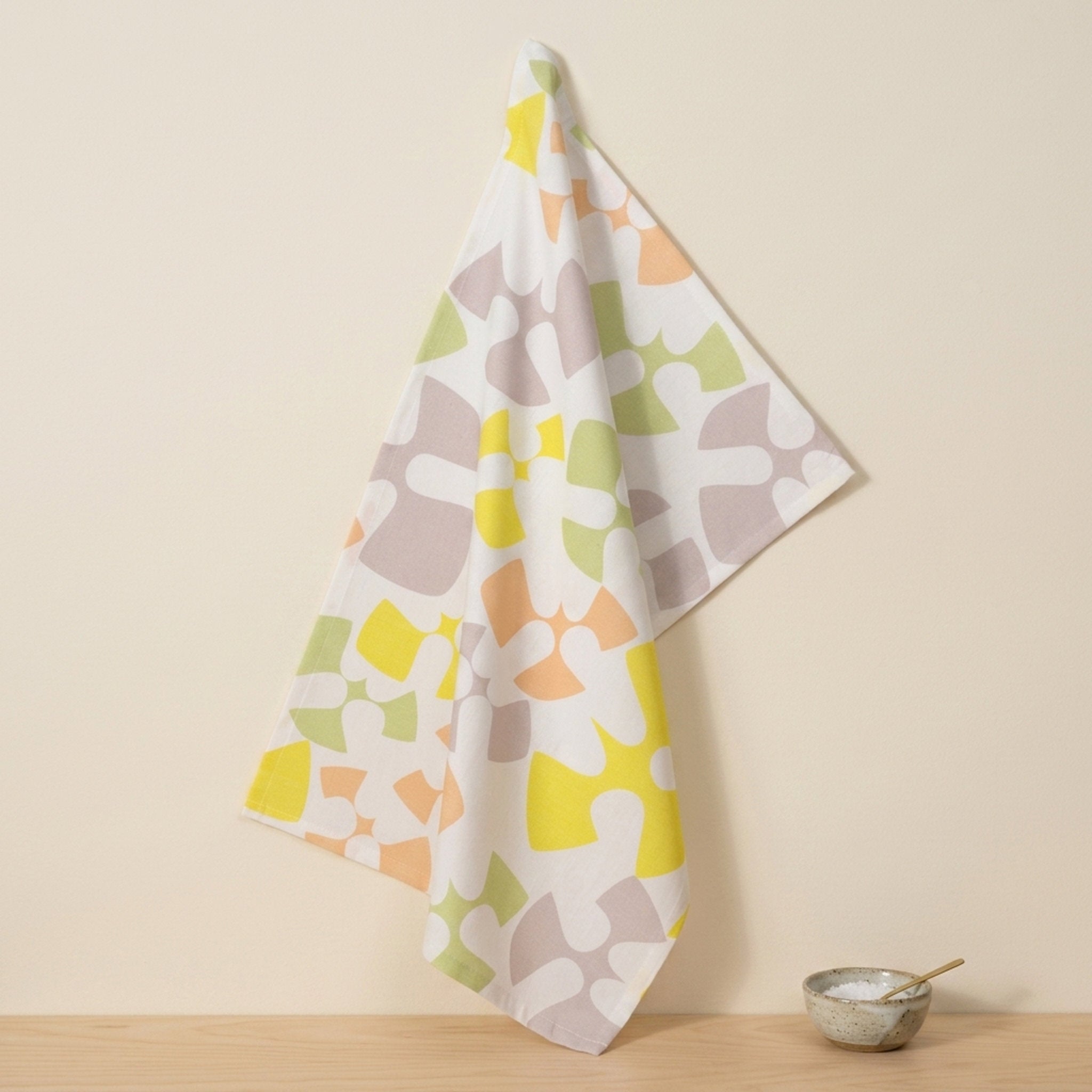A white kitchen towel with a colorful abstract organic pattern of flying birds in yellow, orange, green, and gray, hanging on a wall hook next to a small ceramic bowl on a wooden surface from Hem And Tassel