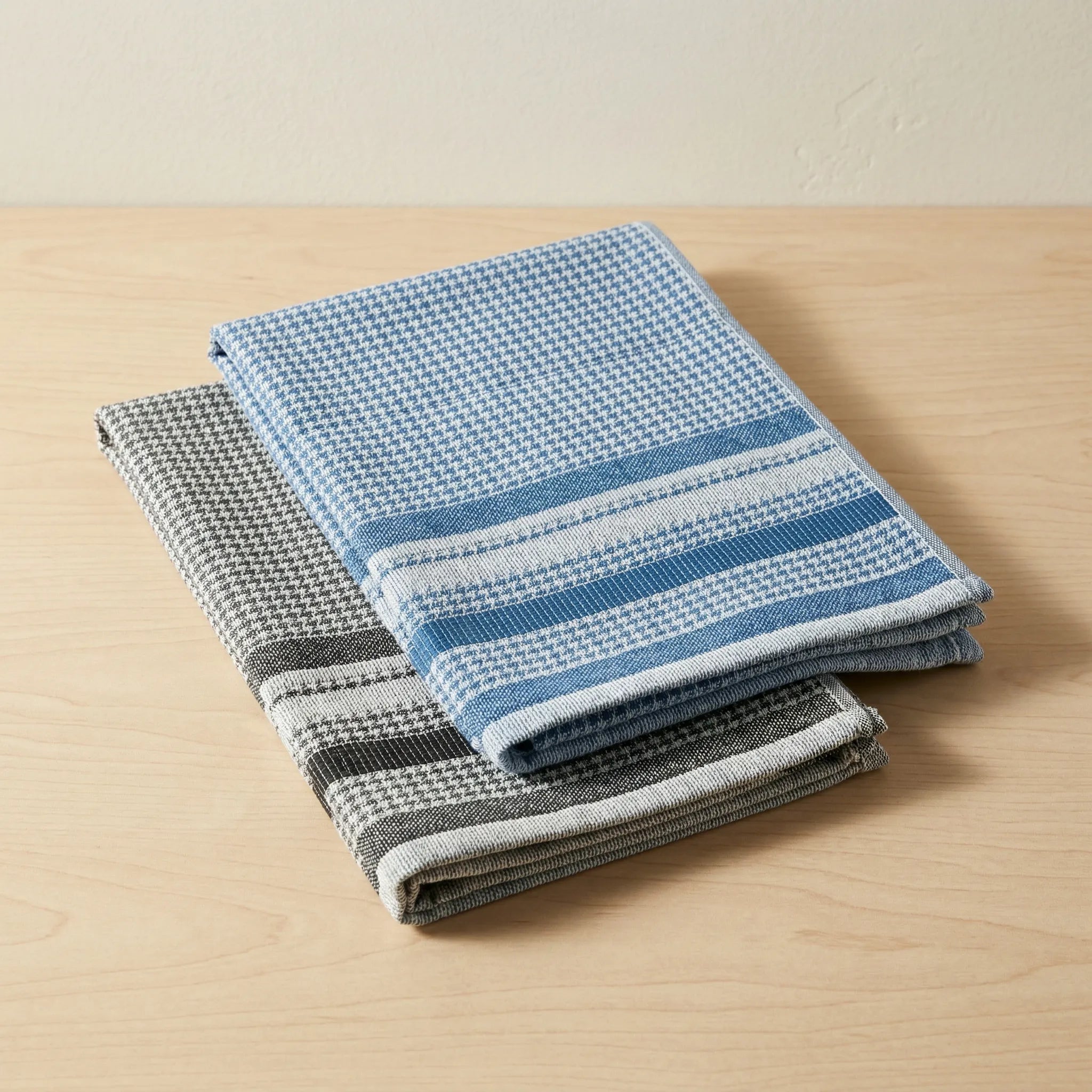Jacquard kitchen towels of 100% cotton with all-over houndstooth pattern - one in blue and one in black, folded and stacked on a light colored wood backdrop from Hem And Tassel 