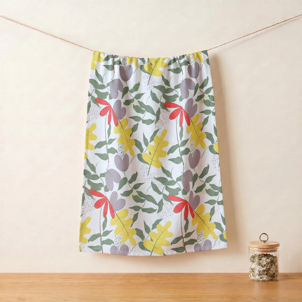 A gathered cotton kitchen towel with a colorful botanical pattern of yellow leaves, red bows, and gray flowers on a white background, hanging from a string against a beige wall from Hem And Tassel 
