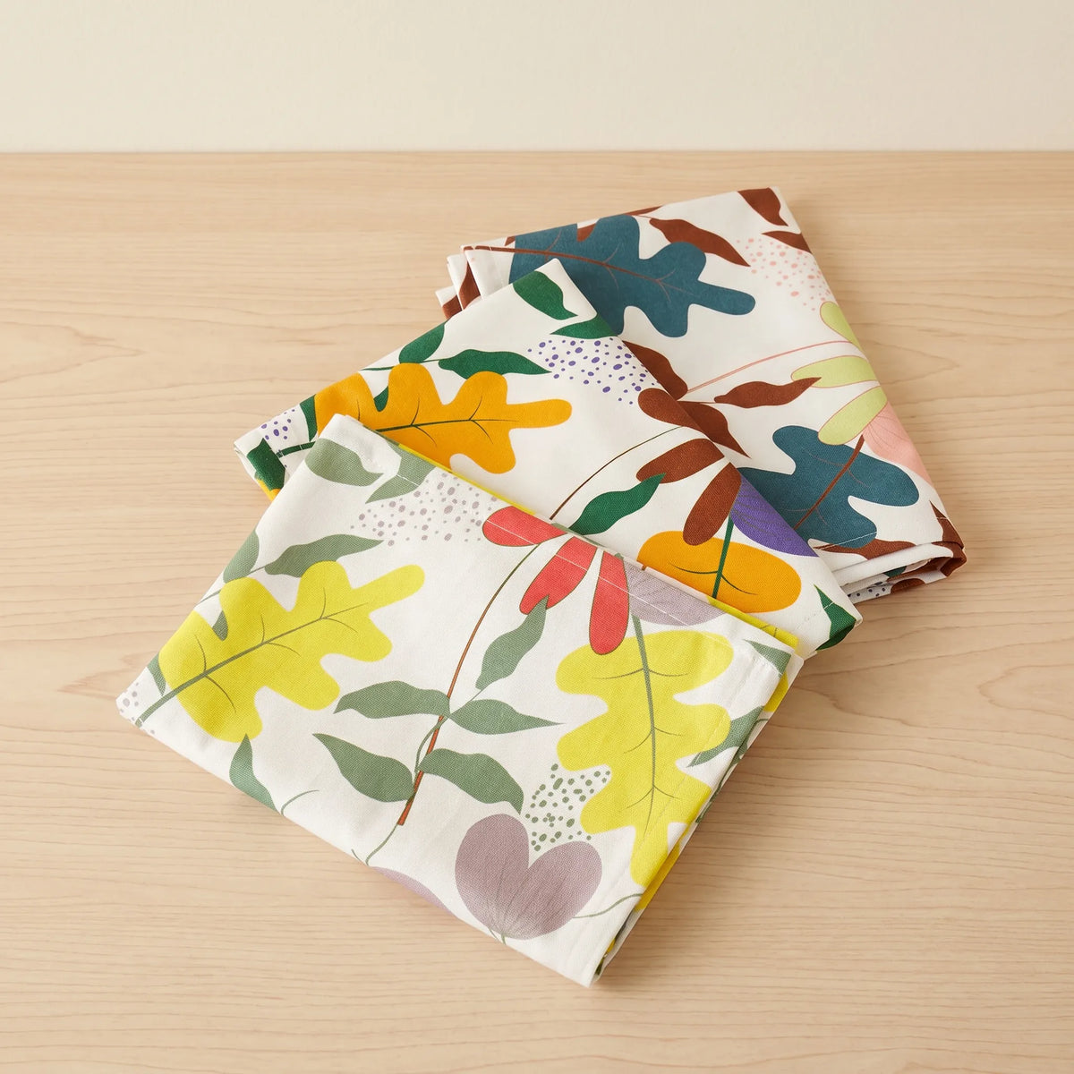 A stack of folded cotton kitchen towels with a colorful autumn leaf pattern on a cream background, displayed on a light wood tabletop from Hem And Tassel 