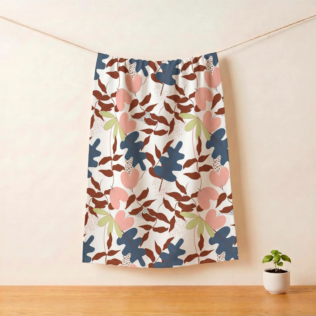 A patterned cotton kitchen towelhanging from a rope against a beige wall, with a small potted plant on a wooden surface below. The kitchen towel features a colorful botanical pattern of  with pink hearts, blue and brown leaves, and light green leaves on a white background from Hem And Tassel 
