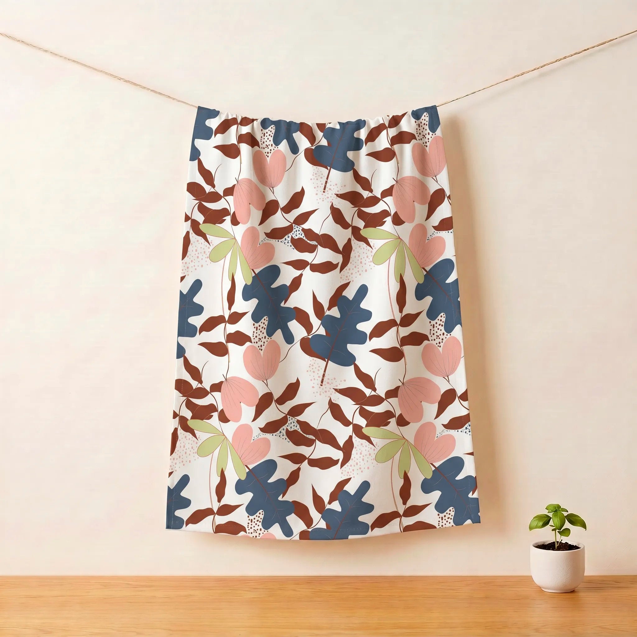 A patterned cotton kitchen towelhanging from a rope against a beige wall, with a small potted plant on a wooden surface below. The kitchen towel features a colorful botanical pattern of  with pink hearts, blue and brown leaves, and light green leaves on a white background from Hem And Tassel 