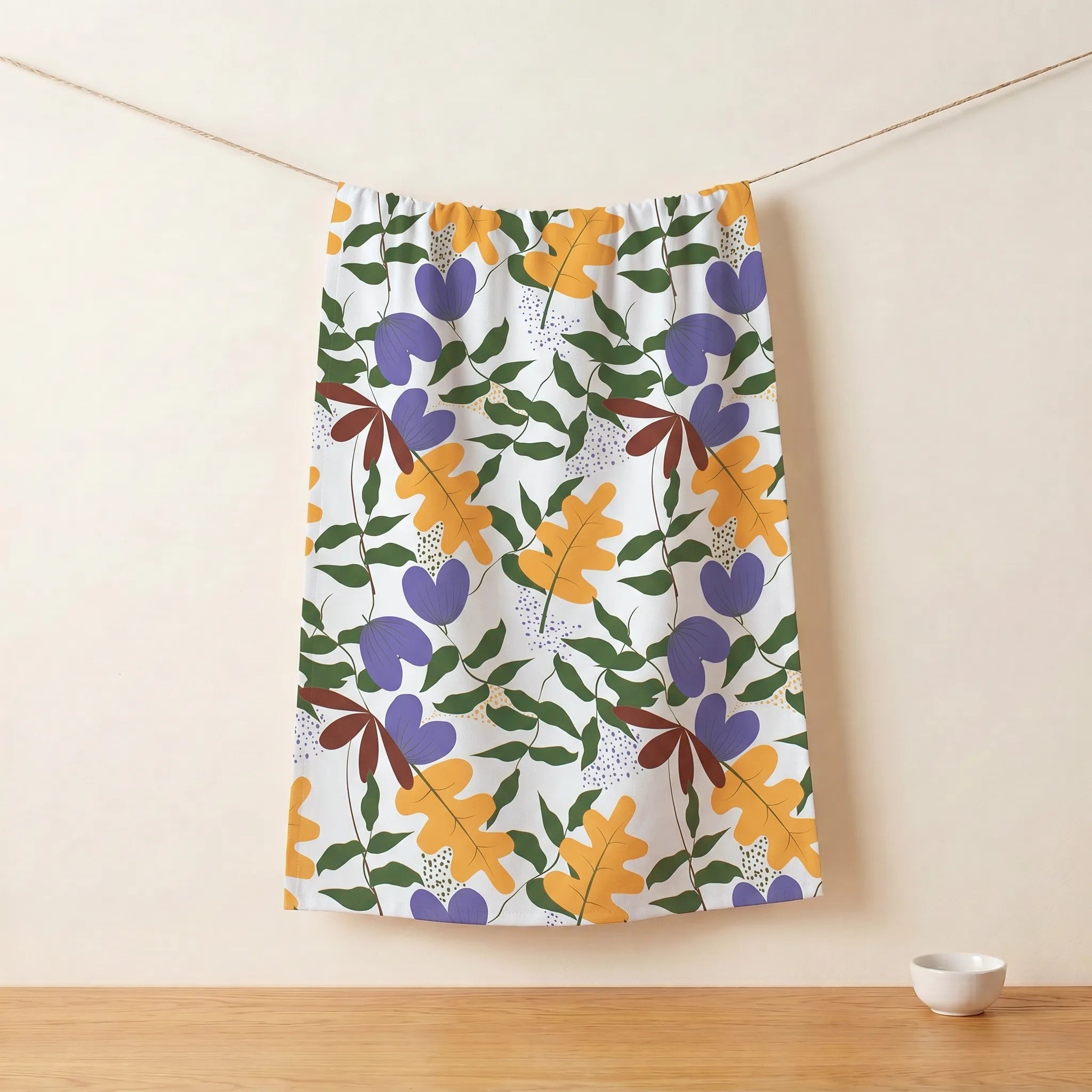 A white cotton kitchen towel featuring a colorful botanical pattern of orange, purple, green, and brown leaves, hanging from a string against a beige wall with a wooden floor from Hem And Tassel 