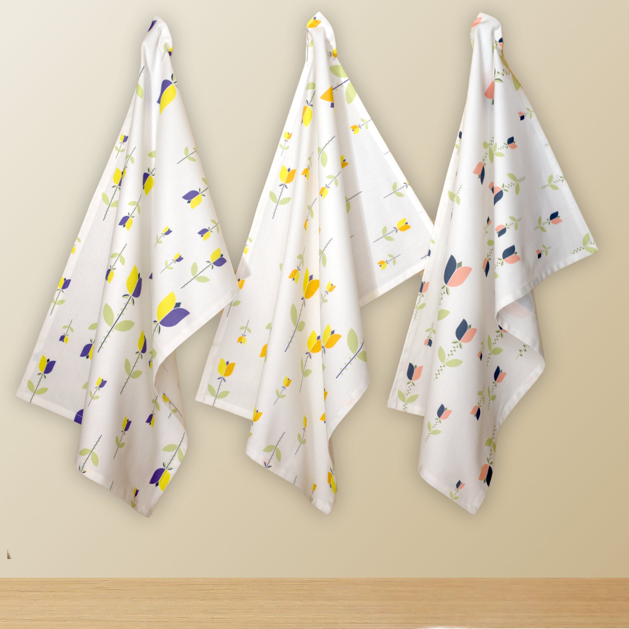 Three white kitchen towels with a colorful floral pattern hang on hooks against a beige wall from Hem And Tassel