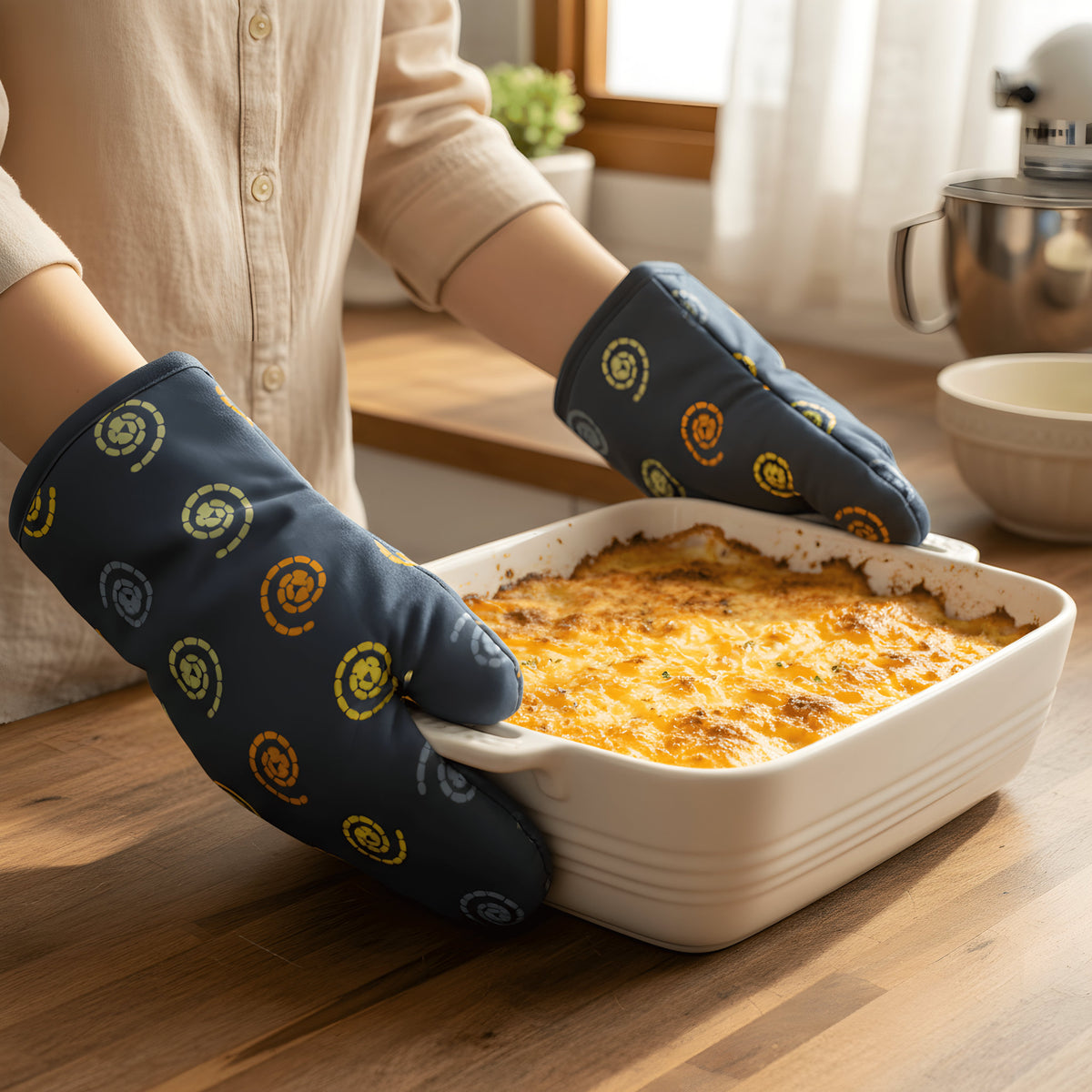 A person wearing dark oil blue oven mitts with yellow and orange spiral patterns holds a white ceramic baking dish filled with a cheesy baked casserole on a wooden kitchen counter from Hem And Tassel 