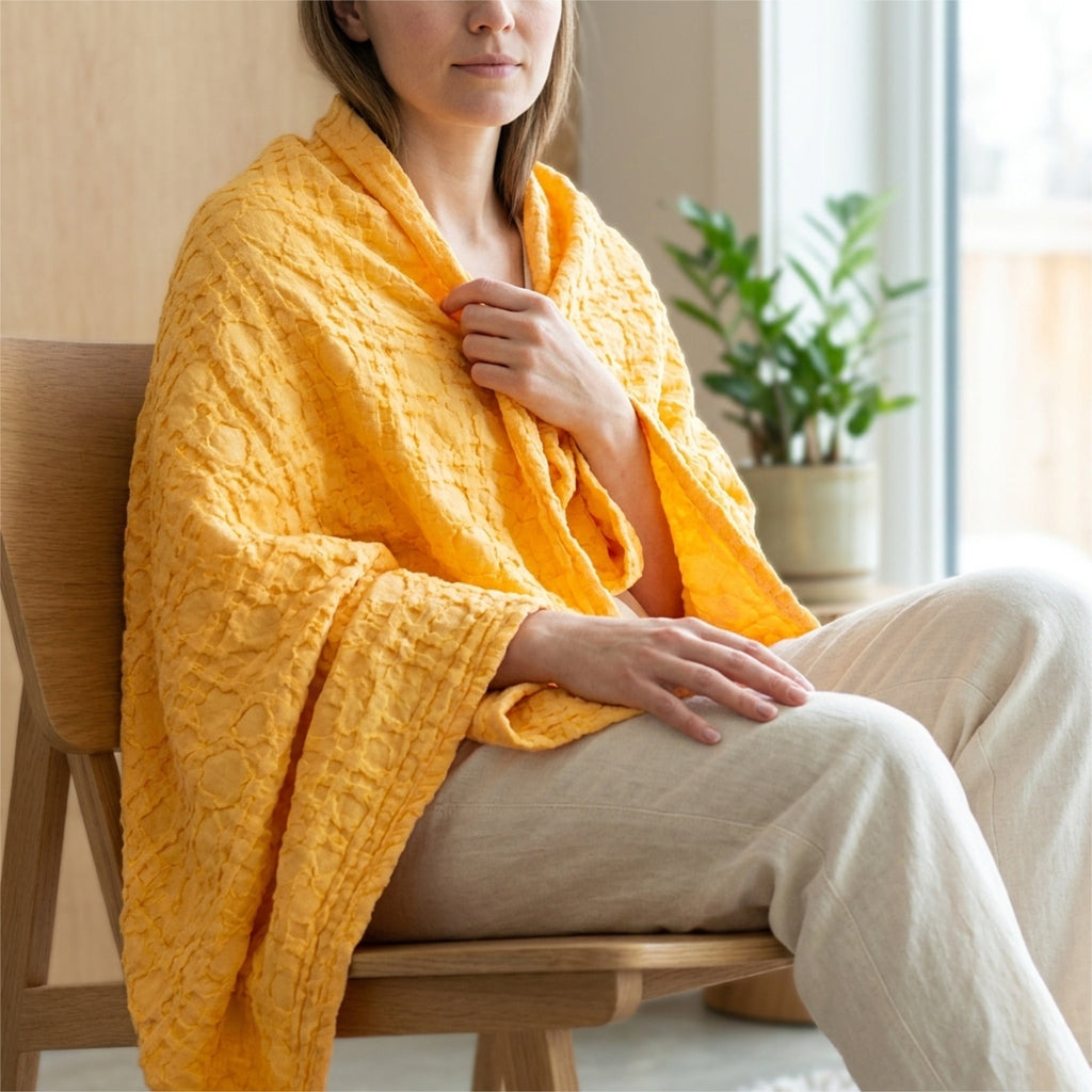 A woman seated on a wooden chair indoors, wrapped in a box waffle mild mango colored woven throw blanket hemmed on four sides. She is wearing light beige pants, with a potted plant and soft window light in the background from Hem And Tassel 