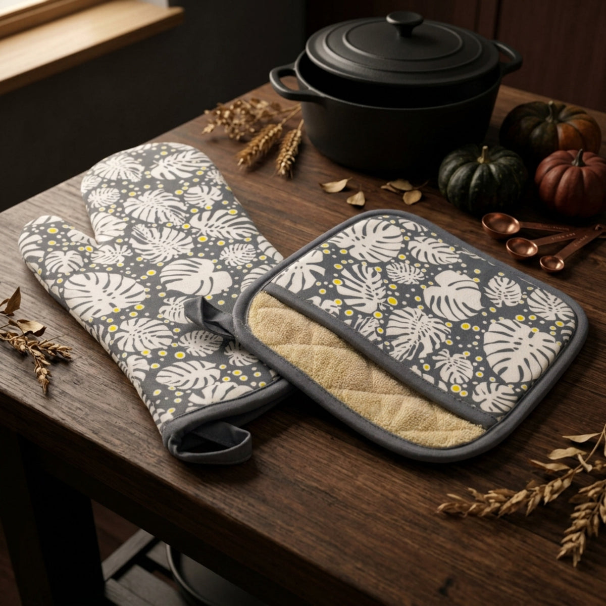 Two oven mitts and a pot holder with a gray and white tropical leaf pattern and small yellow dots, displayed on a dark wooden table with autumn props including dried wheat, pumpkins, and a black pot.