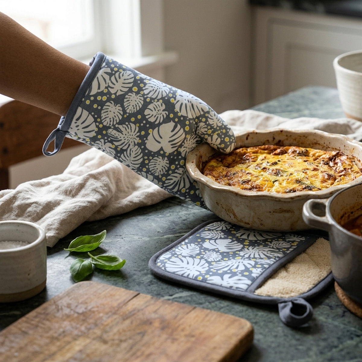 A person wearing a gray kitchen oven mitt with a white tropical monstera leaf pattern and yellow dots, lifting a baked pie from a rustic countertop. A matching trivet and second oven mitt are also visible on the counter in a kitchen setting from Hem And Tassel 