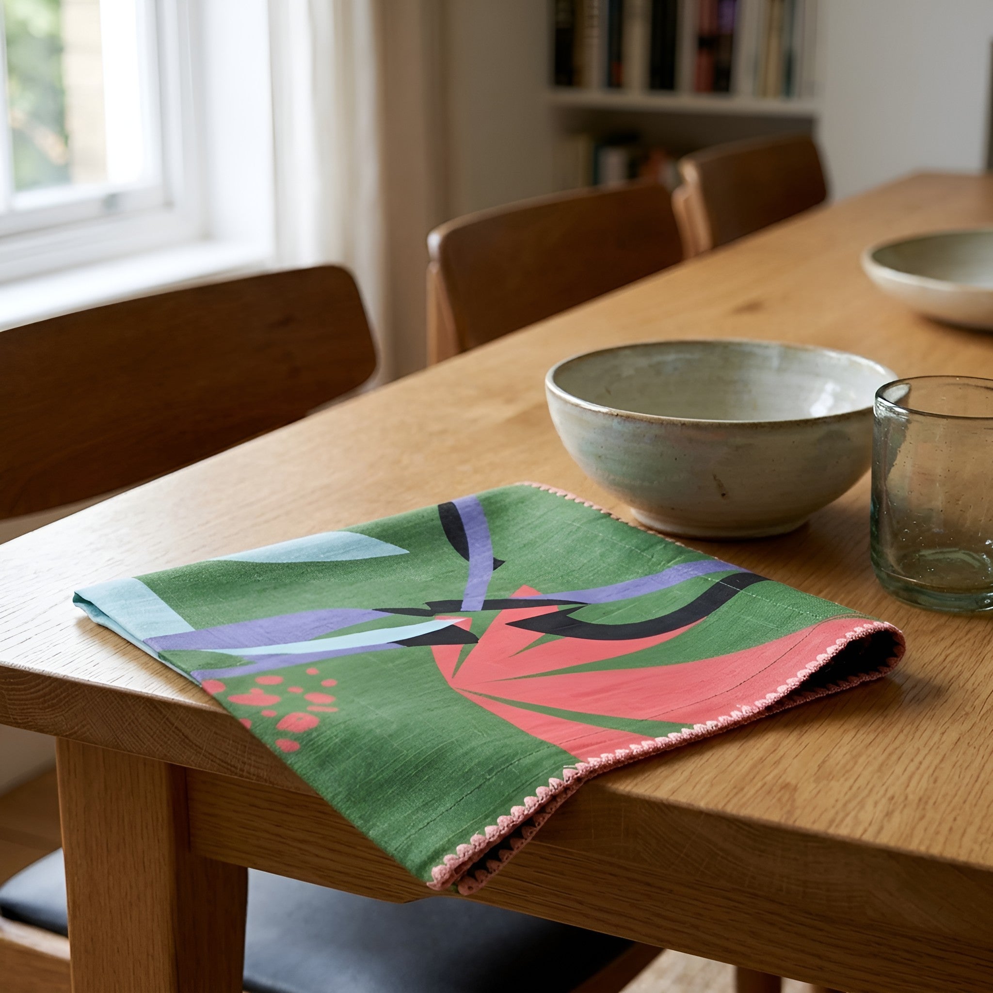 A green dinner napkin with a colorful abstract floral pattern in pink, purple, and black, styled on a wooden dining table next to ceramic bowls and a glass from Hem And Tassel 