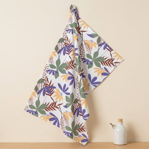 A cotton kitchen towel with a colorful botanical leaf pattern in buff, purple, green, and brown on a white background, hanging against a beige wall above a wooden surface with a small bottle of oil from Hem And Tassel