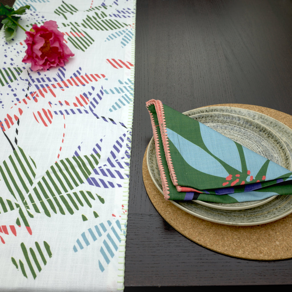 Colorful Pollen up table runner with floral pattern next to a stack of plates and napkins on a dark surface from Hem And Tassel 